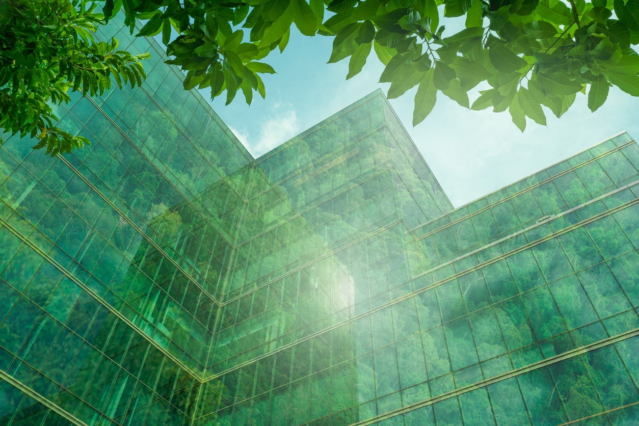 An upward view of a green stretch code building showcasing energy-efficient features can be seen in a modern city. This sustainable glass office building incorporates a tree to reduce heat and utilizes energy-efficient lighting. FreMarq Innovations